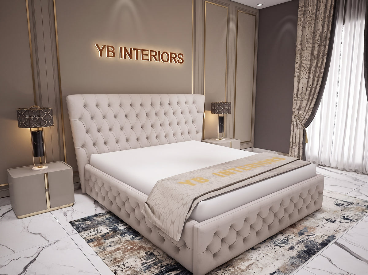 A luxury upholstered bed with a tufted headboard and matching footboard, styled in a modern bedroom setting. The bed is dressed in white bedding with a beige throw blanket featuring gold lettering.
