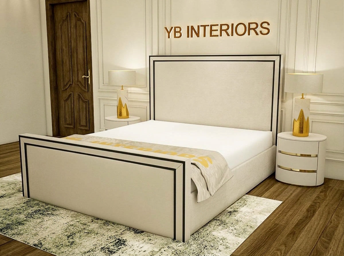 A modern upholstered bed frame in a warm beige fabric with black trim, shown in a styled bedroom setting. The bed is dressed in white bedding with a gold patterned throw.