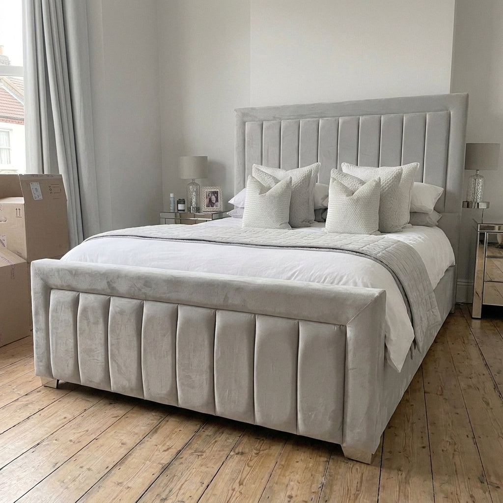 A light gray upholstered bed with a tall channel-tufted headboard and matching footboard, styled in a bedroom with white bedding, pillows, wooden floors, and bedside tables.