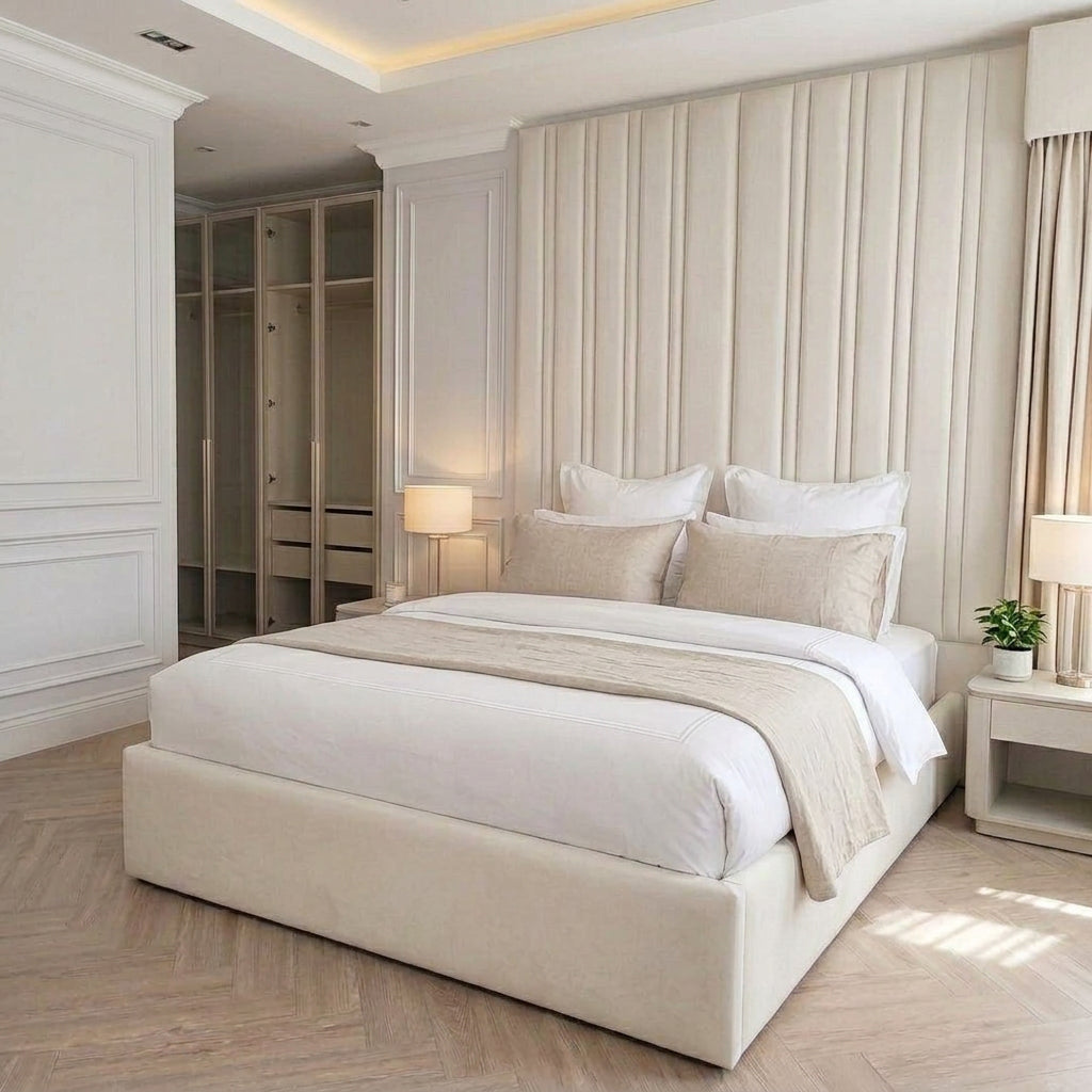 A modern bedroom featuring a large upholstered platform bed in cream beige, set against a matching pleated headboard. The room has light wood flooring, white walls with classic molding, and a built-in wooden wardrobe in the background.