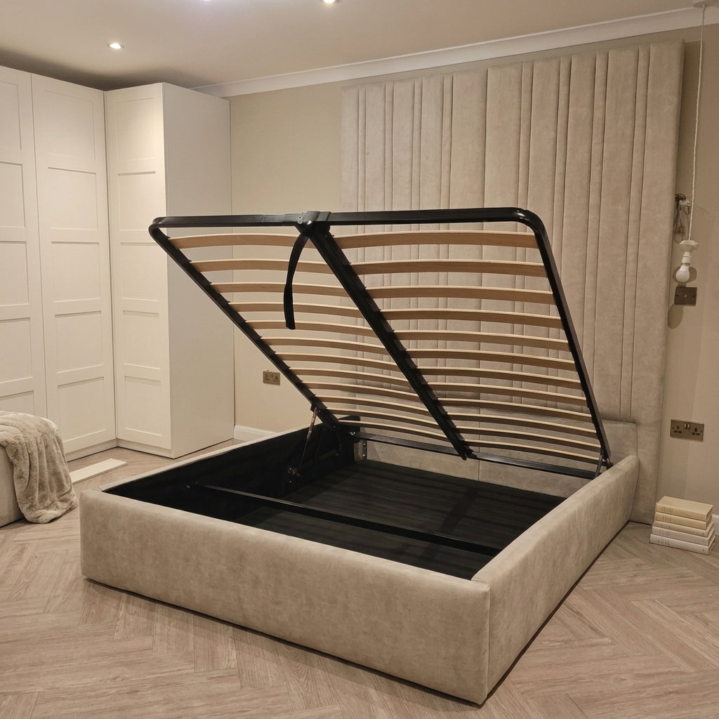 A beige upholstered lift-up platform bed with its black metal support frame open, showing the storage compartment underneath, set in a modern bedroom with beige curtains and light wood flooring.