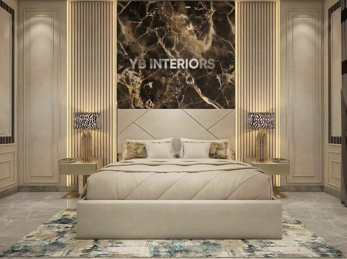 A luxury modern bedroom featuring a large beige upholstered bed with a matching footboard, flanked by gold-toned nightstands and lamps. The background is dominated by a large black and gold marble wall panel with vertical gold slats, and the brand name 'YB INTERIORS' is printed on the panel.