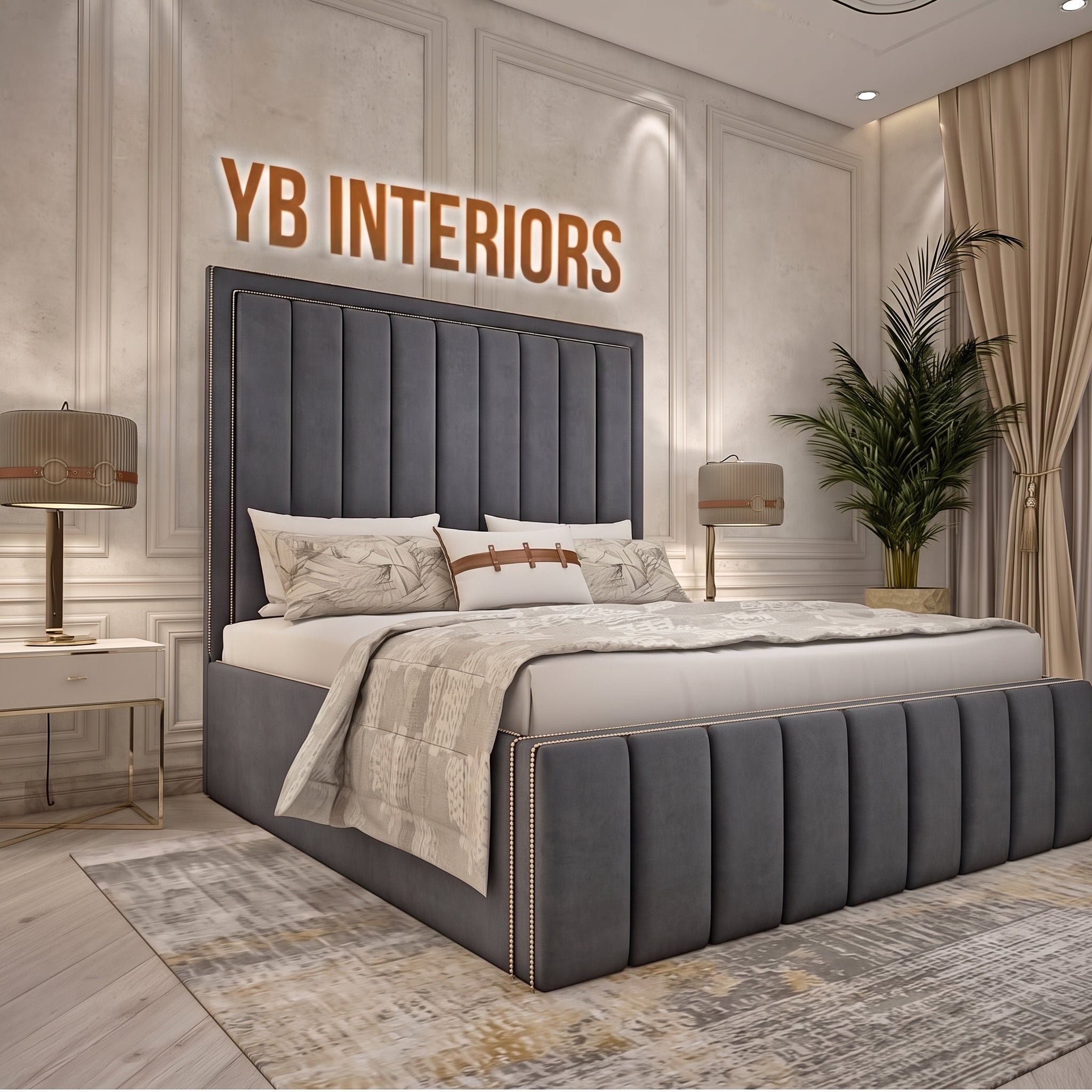 A modern upholstered bed with a tall channel-tufted headboard and matching footboard in dark gray velvet, styled in a luxury bedroom. The room features beige walls with decorative molding, a large wooden 'YB INTERIORS' sign on the wall, a patterned beige rug, bedside tables with lamps, and a potted palm.