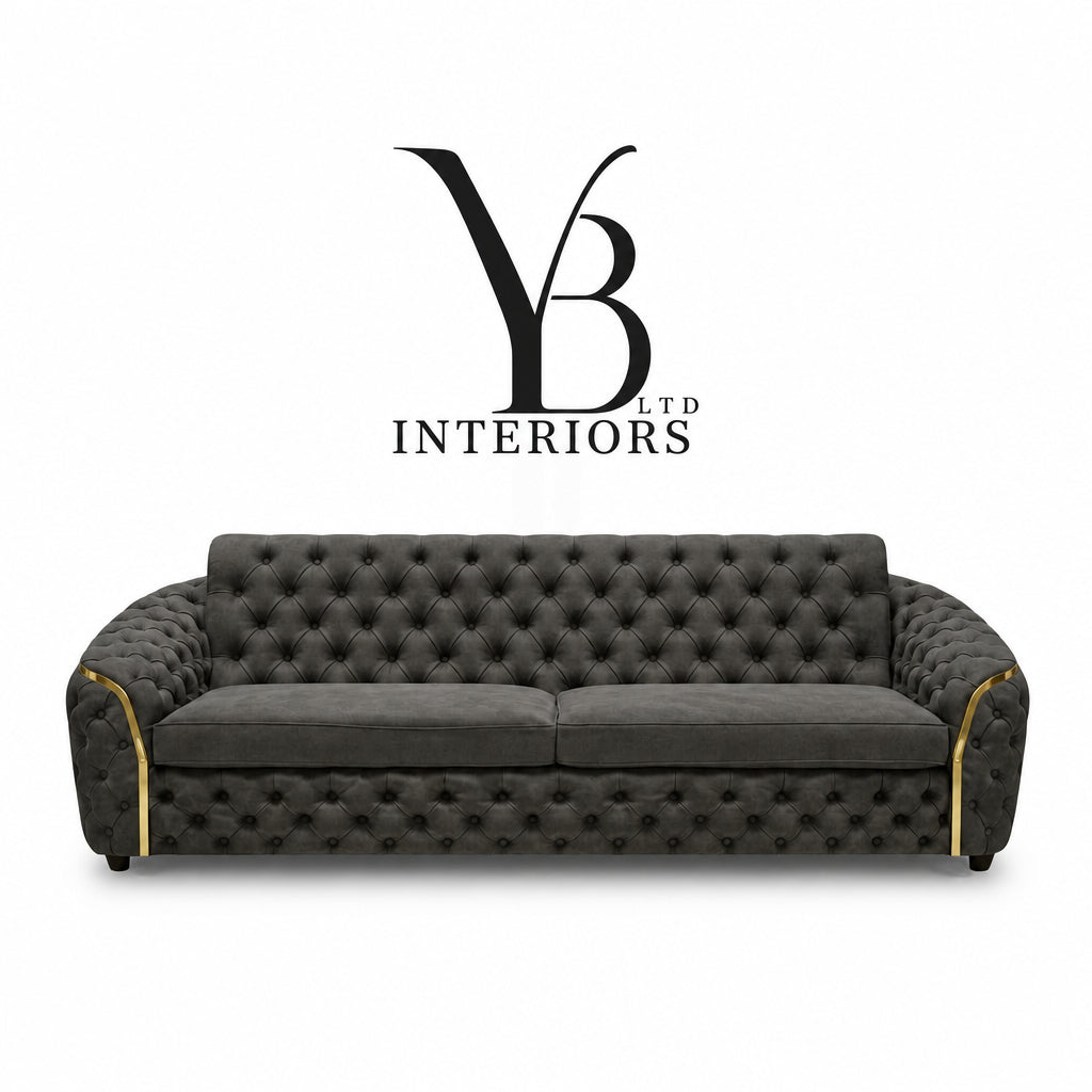 A dark gray tufted velvet sofa with gold trim on the armrests, shown against a white background with the 'YB Interiors' logo above it.