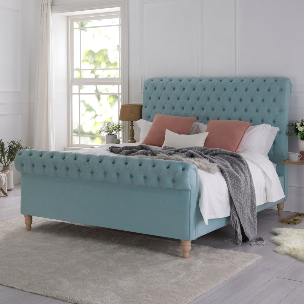 A light blue tufted velvet bed frame with wooden legs, styled with white and dusty rose bedding in a bright, modern bedroom.