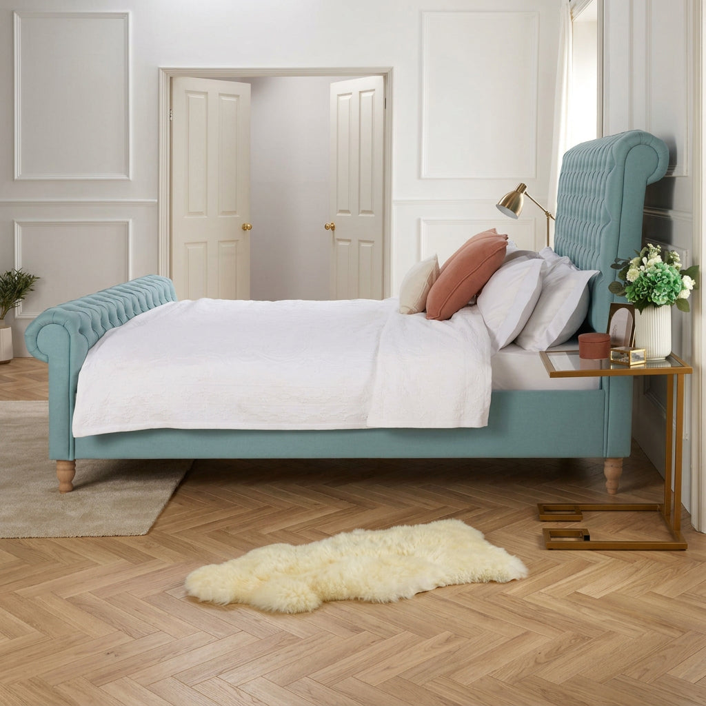 A light blue upholstered bed with a tall tufted headboard and rolled arms, styled with white bedding and accent pillows in a bright bedroom with wood flooring and a small side table.