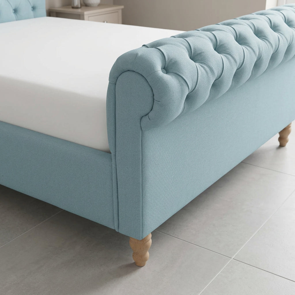 A light blue upholstered bed frame with a tufted headboard and turned wooden legs, shown with a white mattress on a light gray tiled floor.