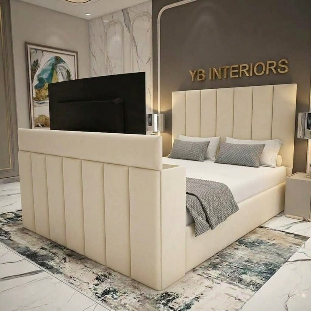A cream upholstered bed frame with a tall segmented headboard and matching footboard, styled in a modern bedroom. A black TV is mounted on the back of the headboard, and the wall behind features gold 'YB INTERIORS' lettering.