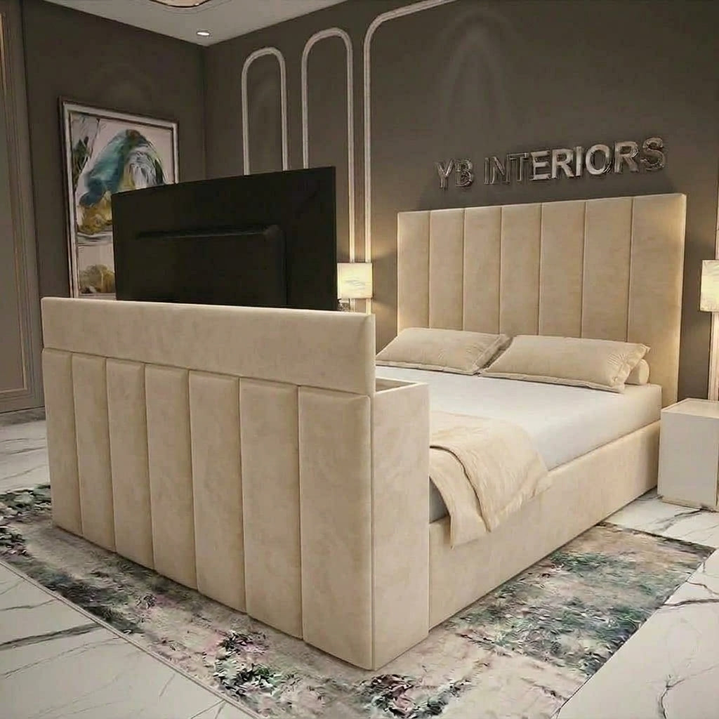 A modern upholstered bed frame in a warm beige fabric, featuring a tall segmented headboard and matching low footboard, styled in a contemporary bedroom with a TV mounted above the bed, bedside tables, lamps, and a patterned rug.