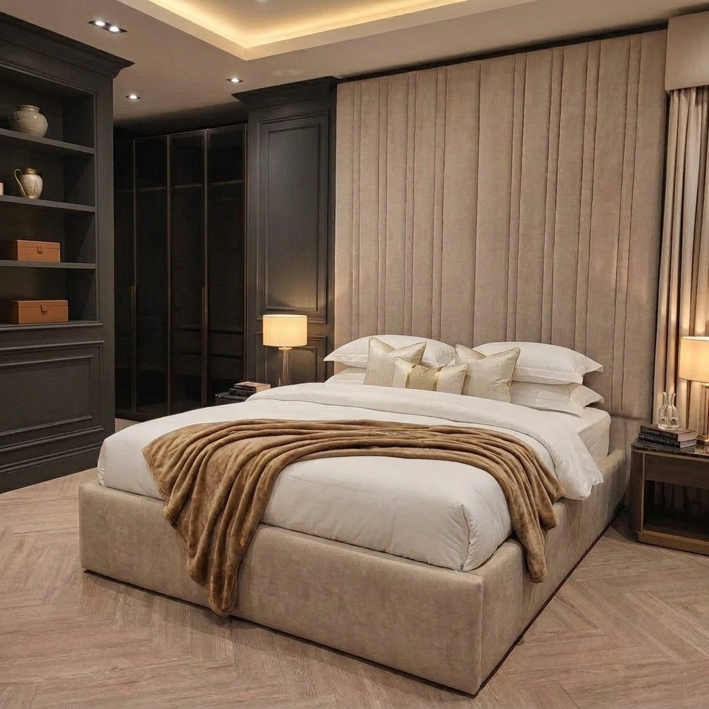 A luxury bedroom featuring a large upholstered platform bed in beige, styled with white bedding and a brown throw blanket. The background is dominated by a tall, pleated beige headboard wall, flanked by dark wood paneling, nightstands, and lamps.