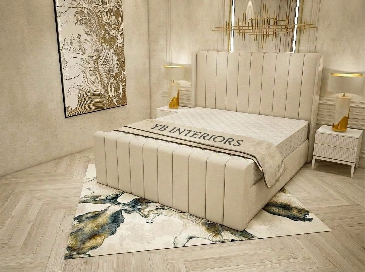 A cream upholstered bed frame with a tall vertical headboard and matching footboard, shown in a styled bedroom setting with a patterned rug, bedside tables, lamps, and wall art. The bedding has 'YB INTERIORS' printed across it.