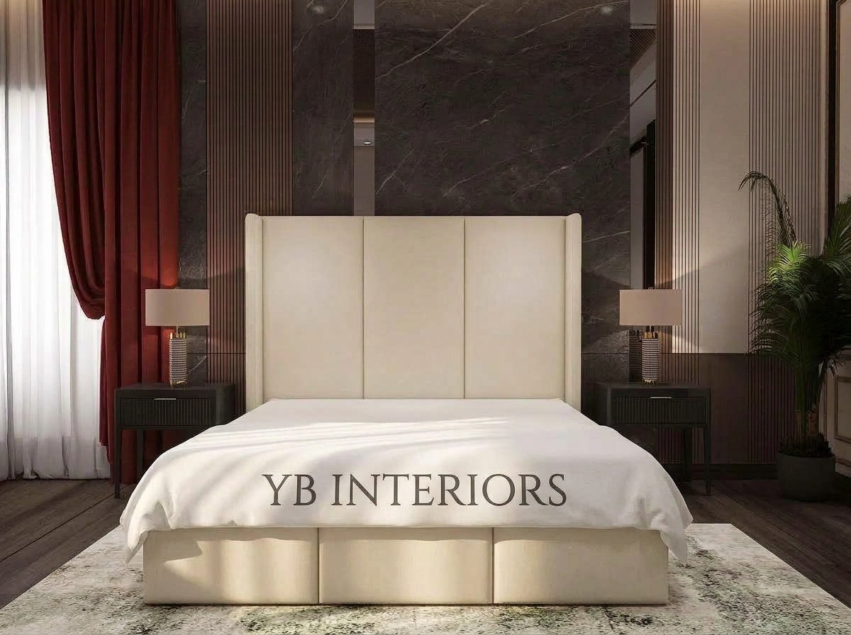 A modern upholstered bed with a tall segmented headboard and matching footboard in a warm cream or beige tone, shown in a styled bedroom. The bed is dressed in white bedding, with the text 'YB INTERIORS' printed across the lower front.