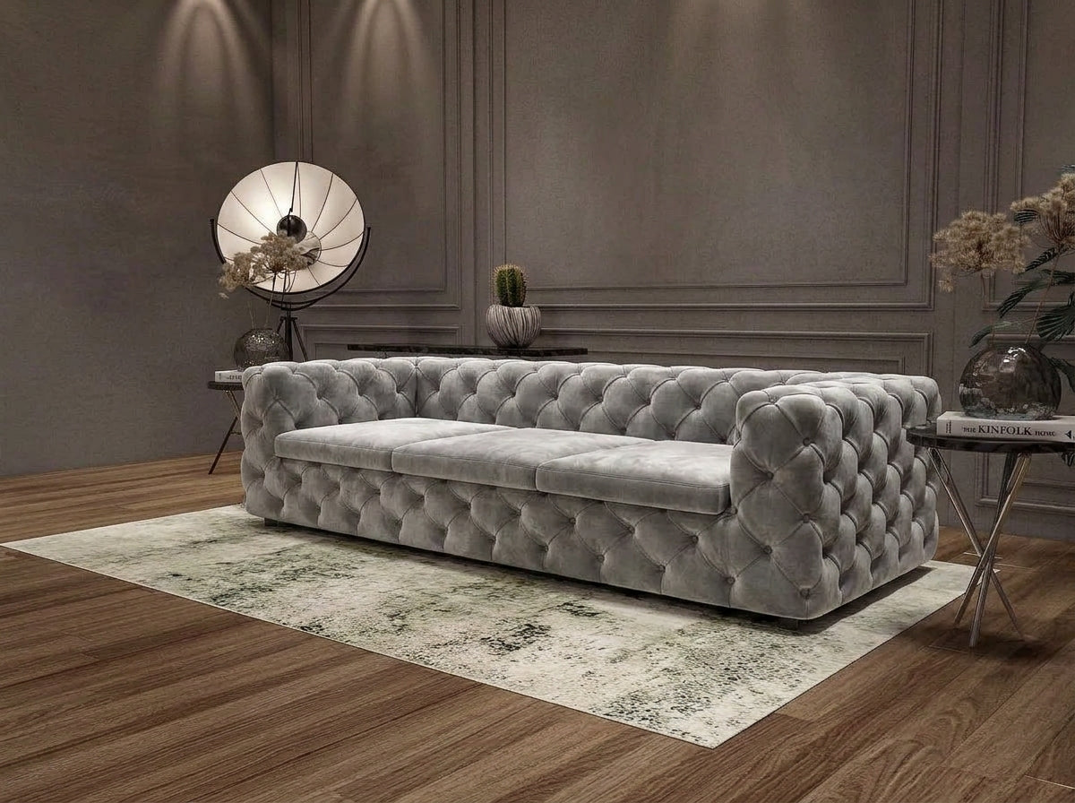 A light gray tufted velvet sofa with a chaise lounge attachment, styled in a modern living room setting on a patterned rug with wood flooring.
