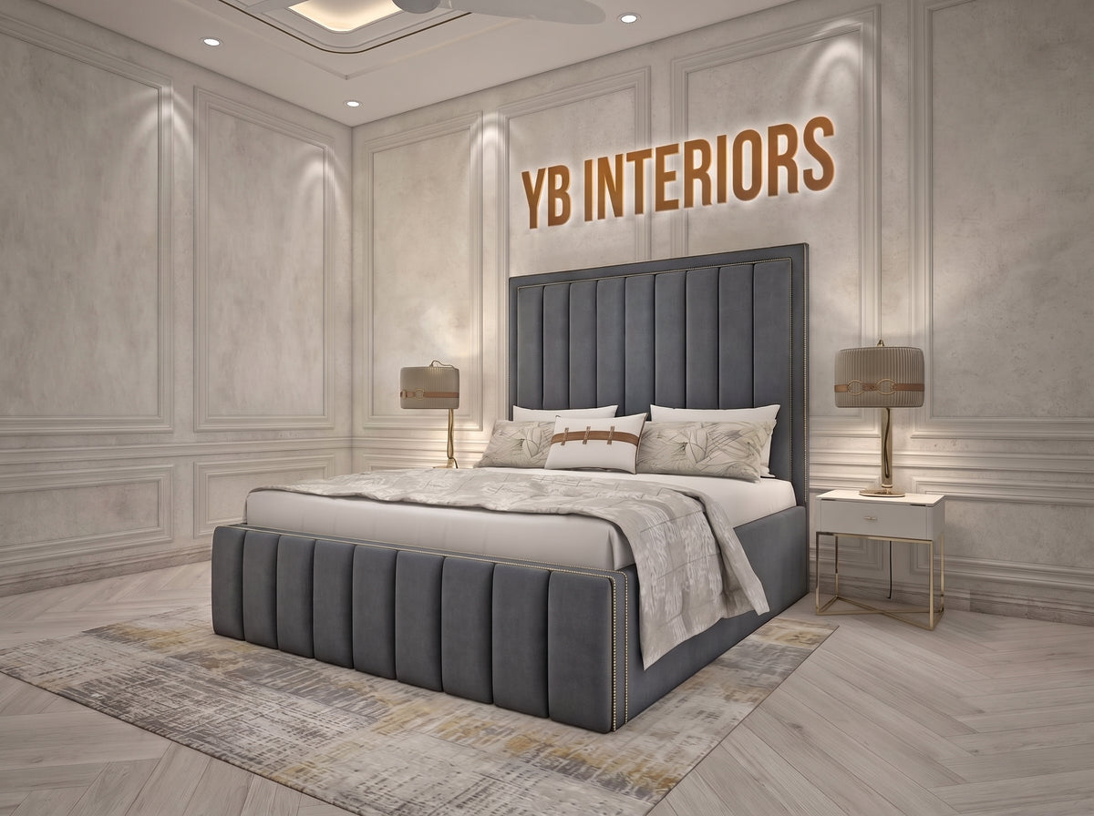 A modern gray tufted platform bed with a tall channel-tufted headboard, styled in a luxury bedroom with light gray paneled walls, gold accent lighting, and a matching gray rug. The text 'YB INTERIORS' is displayed on the wall above the headboard.